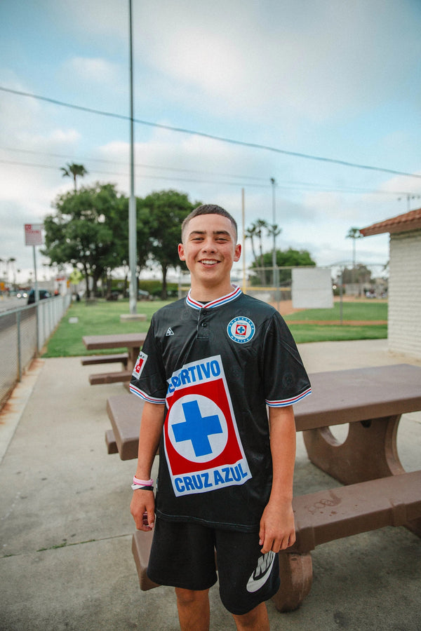 A young man smiling outdoors wearing a black MadStrange Cruz Azul 1997 Champions soccer jersey with a red and blue crest.
